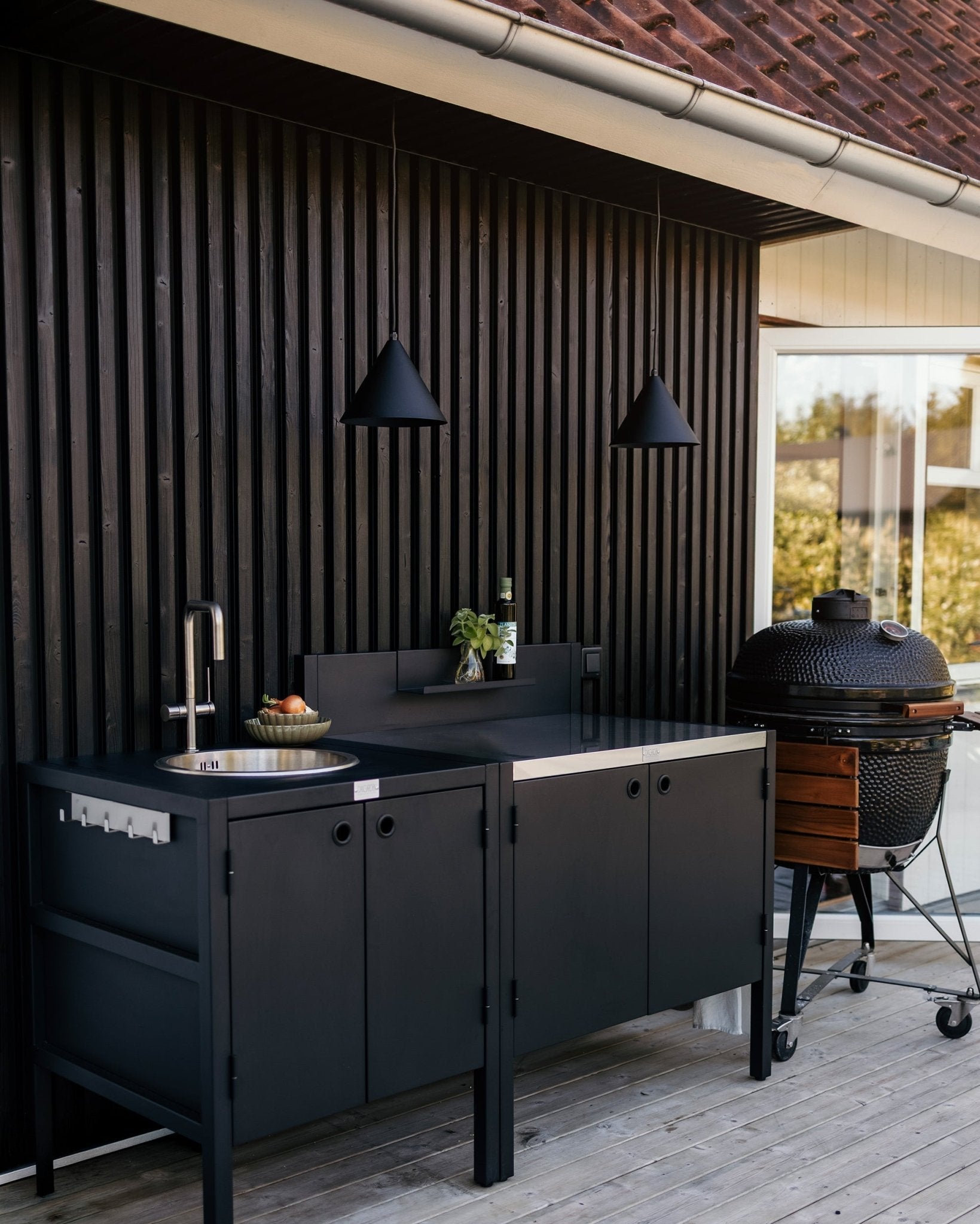 Modern outdoor kitchen with matte black cabinets, a sink, two cone-shaped pendant lights, a grill on the right, and a bottle of wine on the countertop, against a dark wood slatted wall.
